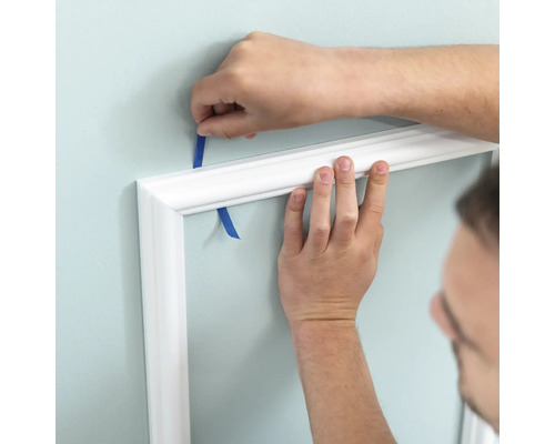 Osoba připevňuje bílou ozdobnou lištu lepicí páskou ke zdi.