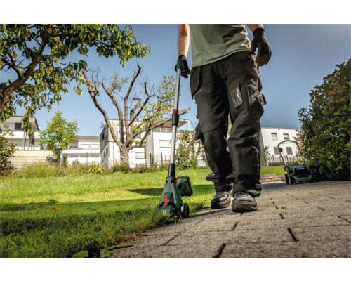 Muž používá akumulátorový vyžínač na zahradě, v pozadí je sekačka na trávu.