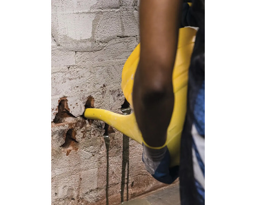 Osoba nalévá tekutinu z konve do poškozené cihlové zdi.