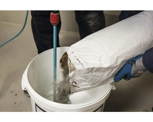 Osoba sype materiál ze sáčku do kbelíku k promíchání.