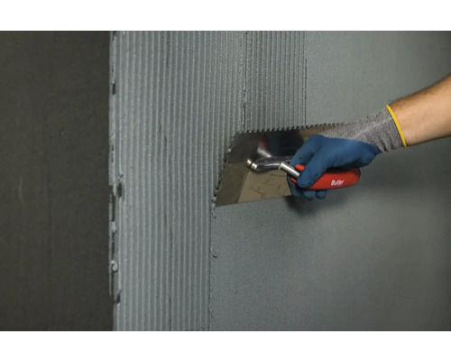 Řemeslník nanáší zubovou stěrkou lepidlo na dlaždice na stěnu.