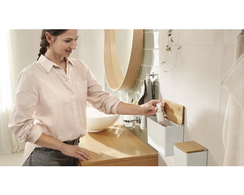 Světlá koupelna s umyvadlem, zrcadlem a ženou, která bere lahvičku z nástěnné police.