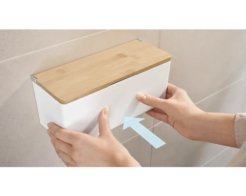 Snímek osoby připevňující bílý box na kosmetické ubrousky s bambusovým víkem na zeď.