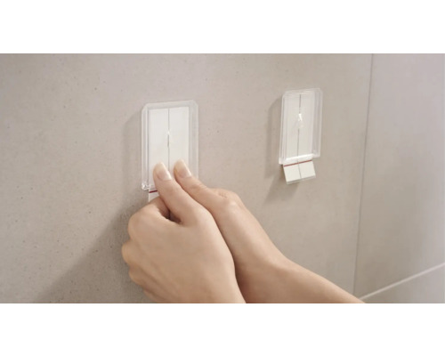 Dva transparentní samolepicí háčky jsou připevněny ke zdi. Jedna ruka táhne za jeden z háčků.