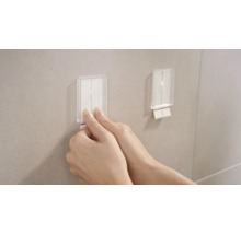 Snímek dvou rukou připevňujících samolepicí háček na zeď.