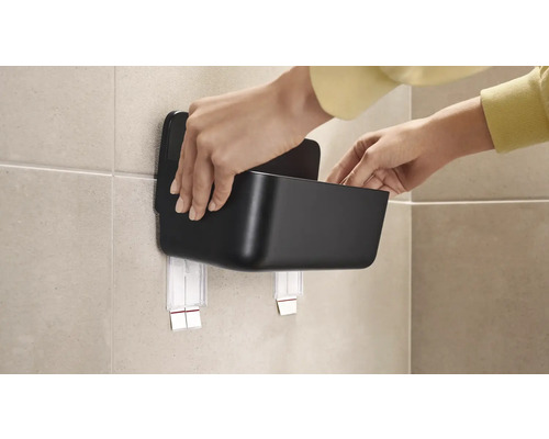 Osoba připevňuje černý úložný box pomocí lepicí upevňovací sady na zeď.