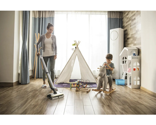 Žena vytírá dřevěnou podlahu v dětském pokoji pomocí akumulátorového mopu, zatímco si dítě hraje na houpacím koni.