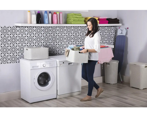 Světlá prádelna s pračkou, sušičkou, košem na prádlo, truhlou na prádlo, policí a ženou nesoucí koš na prádlo.