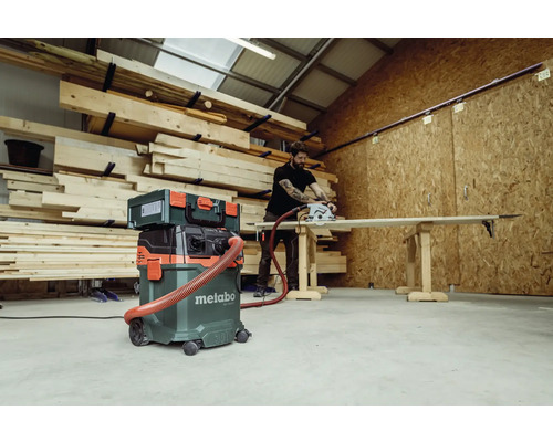 Dílna s uskladněným dřevem, průmyslový vysavač Metabo, kufřík s nářadím a muž řežící dřevo