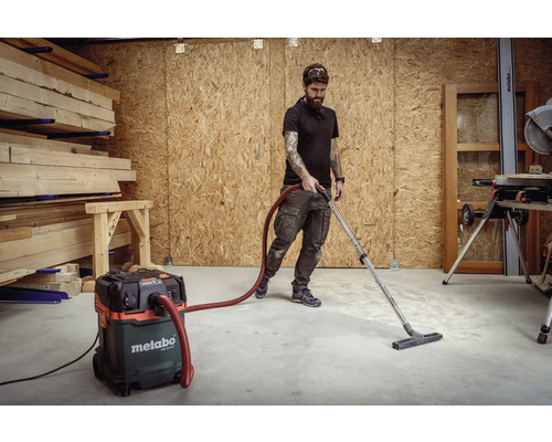 Dílna s mužem, který používá vysavač Metabo k čištění podlahy, se stohy dřeva v pozadí.