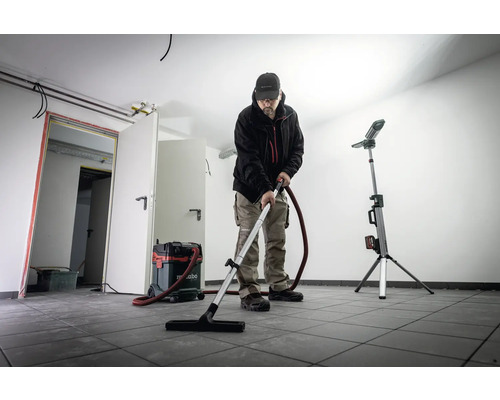 Muž vysává místnost s dlaždicemi pomocí vysavače Metabo, vedle něj stojí stavební reflektor.
