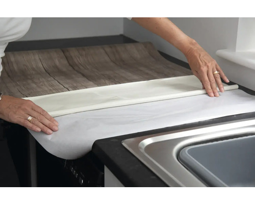 Osoba nanáší nábytkovou fólii s dřevěným vzhledem na kuchyňskou linku.