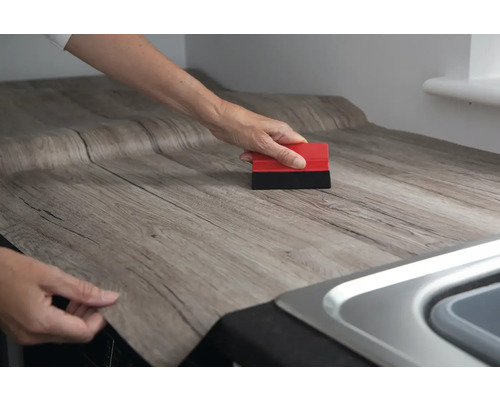 Osoba nanáší dřevěnou dekorativní fólii na pracovní desku pomocí stěrky.