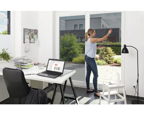 Žena aplikuje protisluneční fólii na okno, v pozadí kancelář