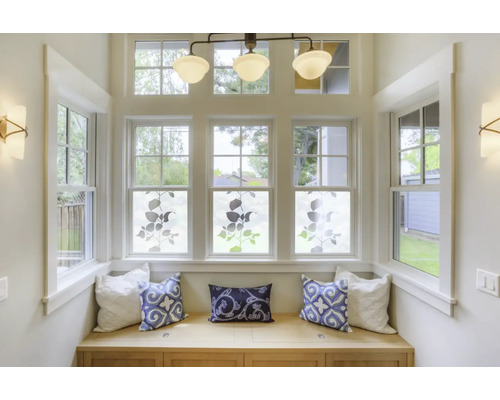 Světlý arkýř s okny zdobenými rostlinnými motivy na okenní fólii, lavice s polštáři, stropní svítidlo a nástěnné lampy