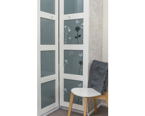 Bílá skříňka s květinovými motivy a skleněnými dvířky vedle židle s šedou dekou