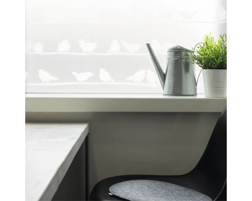 Ozdobné okno s motivem ptáků, konví a rostlinou ve světlé místnosti.