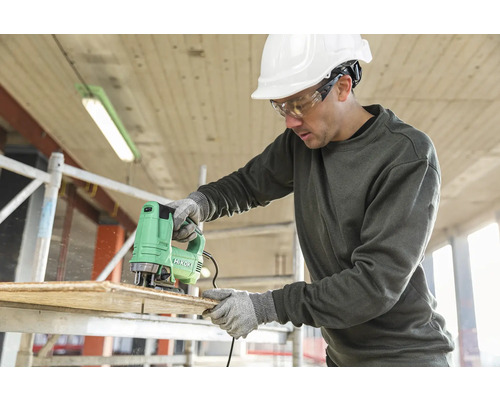 Muž řeže dřevěnou desku přímočarou pilou značky Hikoki a má na sobě ochranné oblečení.