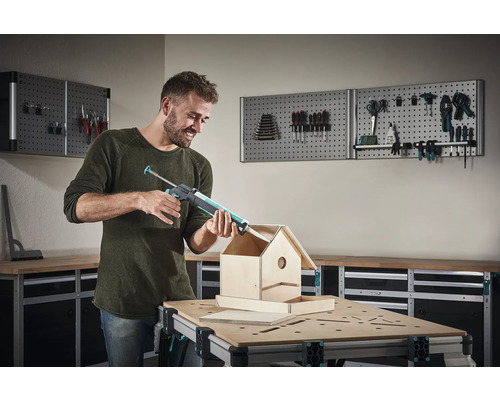 Muž používá pistoli na kartuše ke stavbě ptačí budky na pracovním stole před stěnou s nářadím.