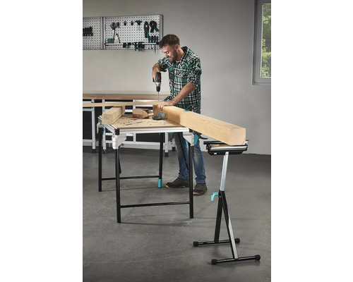 Muž vrtá aku vrtačkou do kusu dřeva na pracovním stole, v pozadí nářadí na stěně s nářadím