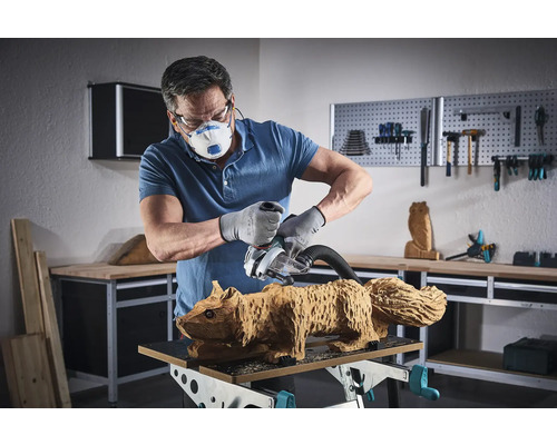Muž brousí brusným strojem dřevěnou figurku veverky. Má na sobě masku, ochranné brýle a rukavice.