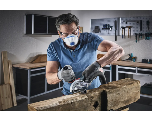 Muž brousí kus dřeva elektrickým nástrojem v dílně, má na sobě ochrannou masku a rukavice.