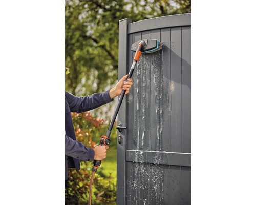 Osoba čistí šedá zahradní vrata mycím kartáčem a zahradní hadicí.