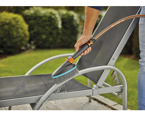 Osoba čistí zahradní lehátko kartáčem a vodní hadicí.