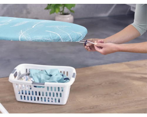 Osoba upevňuje potah na žehlicí prkno s gumičkou, v popředí koš na prádlo