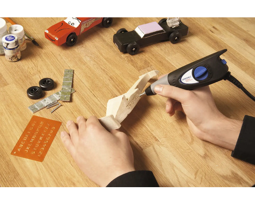 Řemeslná scéna s multifunkčním nástrojem Dremel, dřevěným modelem auta a příslušenstvím na dřevěném stole.