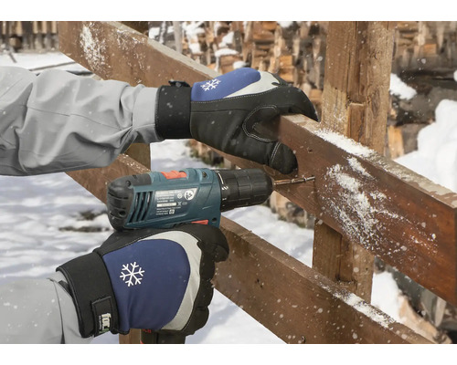 Osoba vrtá otvor do dřevěné desky aku šroubovákem, přičemž má na sobě rukavice