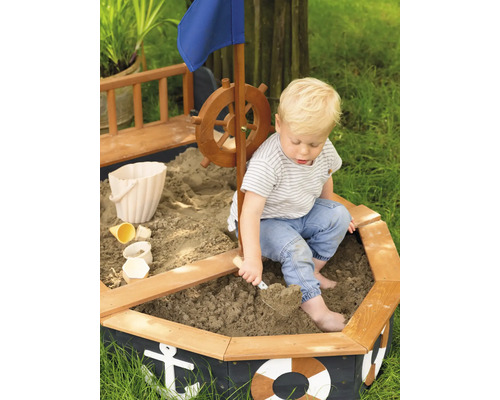 Dítě si hraje v pískovišti ve tvaru lodi s kormidlem, vlajkou a námořnickými motivy.