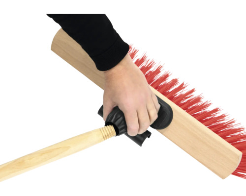 Ruka drží zametací kartáč s dřevěnou rukojetí a červenými štětinami.