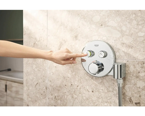 Ruka ovládá sprchovou baterii Grohe na obkládané stěně.