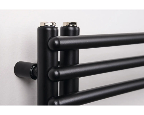 Detailní záběr na černý koupelnový radiátor s horizontálními kulatými trubkami a odvzdušňovacími zátkami nahoře.