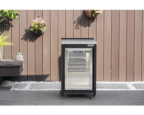 Mobilní venkovní chladnička Tenneker s prosklenými dveřmi a kolečky na terase před dřevěnou stěnou.