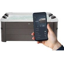 Ruka drží chytrý telefon s aplikací pro ovládání obdélníkové vířivky v šedém dřevěném dekoru, zobrazuje nastavení jako 38 stupňů a bublinky.