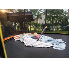 Děti ležící na velké zahradní trampolíně s polštáři a dekami na zahradě vedle dřevěného altánu. Jedno dítě ukazuje na oblohu.