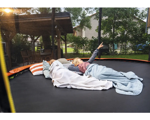 Děti ležící na velké zahradní trampolíně s polštáři a dekami na zahradě vedle dřevěného altánu. Jedno dítě ukazuje na oblohu.
