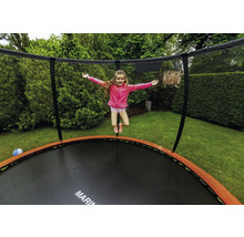 Dítě skáče na kulaté zahradní trampolíně s ochrannou sítí a oranžovým krytem pružin v zelené zahradě.