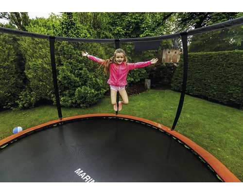 Dítě skáče na kulaté zahradní trampolíně s ochrannou sítí a oranžovým krytem pružin v zelené zahradě.