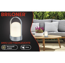 Logo Briloner, stolní LED lampa na akumulátor, teplá bílá 3000 kelvinů, IP44, přenosná s rukojetí, na zahradě na dřevěném stole.