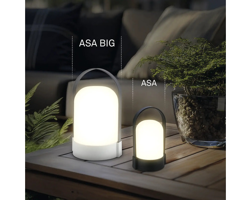 Dvě různě velké přenosné LED stolní lampy ASA BIG a ASA na dřevěném stole venku s rostlinami a čalouněným nábytkem.