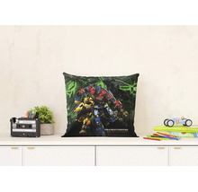 Čtvercový dekorativní polštář s motivem Transformers Probuzení monster na komodě. Světlý pokoj obsahuje rostlinu a hračky.