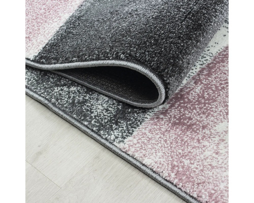 Detailní záběr na koberec s krátkým vlasem, geometrickým vzorem v šedé, růžové a bílé barvě a obšitým okrajem.