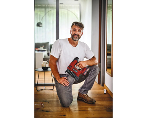 Řemeslník klečí na dřevěné podlaze a drží akumulátorovou sponkovačku Einhell v moderním interiéru.