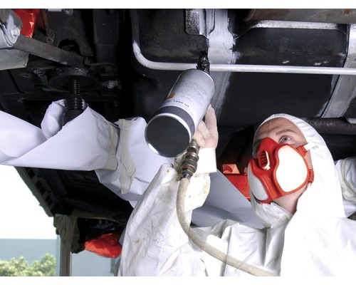 Osoba v ochranném obleku nosí respirátor a stříká ochranu podvozku na zvednuté auto.