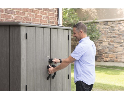 Muž otevírá šedý zahradní box na nářadí s madly a zámkem před cihlovou zdí.