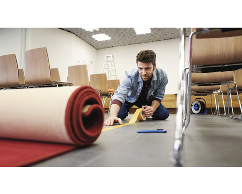 Muž pokládá koberec s lepicí páskou v místnosti se židlemi.