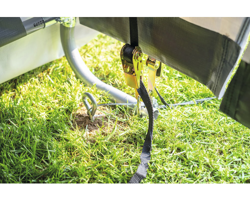 Detailní záběr plachty upevněné k zemi pomocí ráčnového popruhu a háku. Zem je pokrytá trávou.